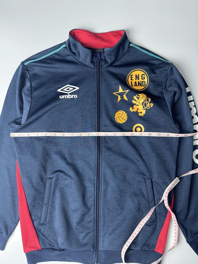 Umbro England Track Jacket Navy Blue (Category: Coats, Jackets and Outerwear) located in Cavite. Submitted by dracokidstuff (Listing ID 1465085774, Image 2). Description: - Brand: Umbro England - Color: Navy blue with red inner collar/panels, teal piping, and white/gold accents - Design: Full zip, dual front pockets, embroidered Umbro logo, gold England crest, and vertical Umbro text on the left sleeve - Fit/Measurements: Width ~20.5" across, Len