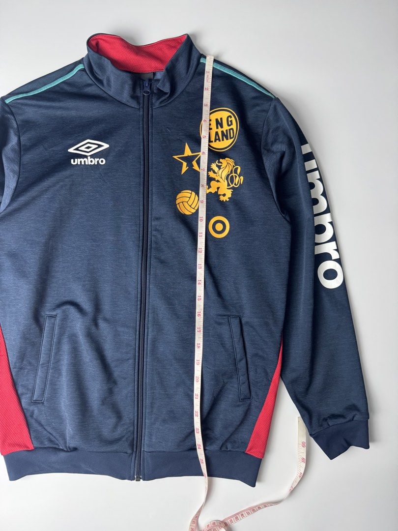Umbro England Track Jacket Navy Blue (Category: Coats, Jackets and Outerwear) located in Cavite. Submitted by dracokidstuff (Listing ID 1465085774, Image 3). Description: - Brand: Umbro England - Color: Navy blue with red inner collar/panels, teal piping, and white/gold accents - Design: Full zip, dual front pockets, embroidered Umbro logo, gold England crest, and vertical Umbro text on the left sleeve - Fit/Measurements: Width ~20.5" across, Len