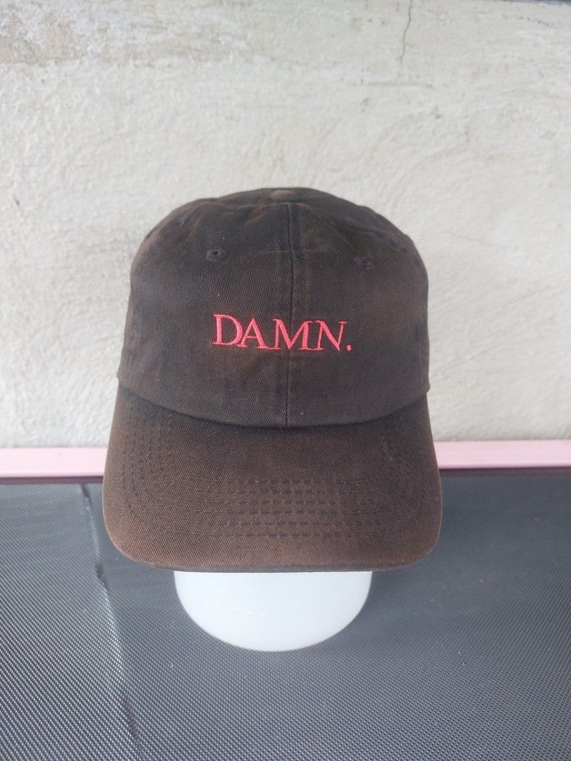 Brown DAMN. Baseball Cap (Category: Caps & Hats) located in Magarao. Submitted by hotdogsandwich.flower (Listing ID 1459600724, Image 1). Description: - Brown baseball cap - Features 'DAMN.' embroidered in red on the front - Adjustable strap at the back for a custom fit - Shows some signs of wear, particularly on the inner sweatband - Still has plenty of life left for everyday use - Perfect for casual outfits.