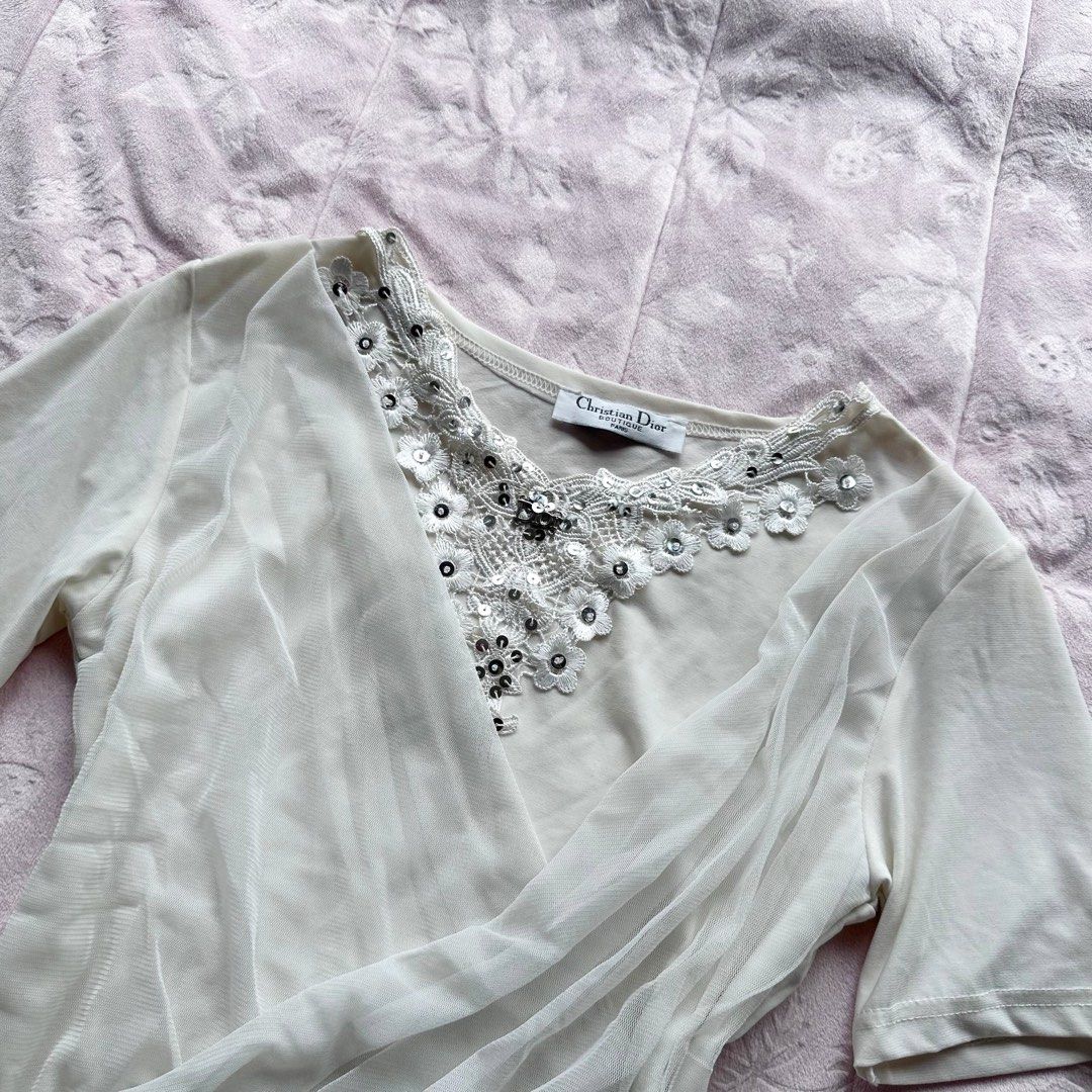 CHRISTIAN DIOR Couture Wrap Top (Category: Blouses) located in San Fernando. Submitted by rianne_von (Listing ID 1459619055, Image 3). Description: W: 15 in L: 20.5 in info: ✧ ivory wrap-style top w/ sheer mesh overlay + draped side detailing ✧ ornate floral lace neckline w/ sequins, beads + Dior logo embellishment ✧ very good thrifted condition; minimal faded stain observed, otherwise no major flaws ✧ color may vary slightly due to lighting.