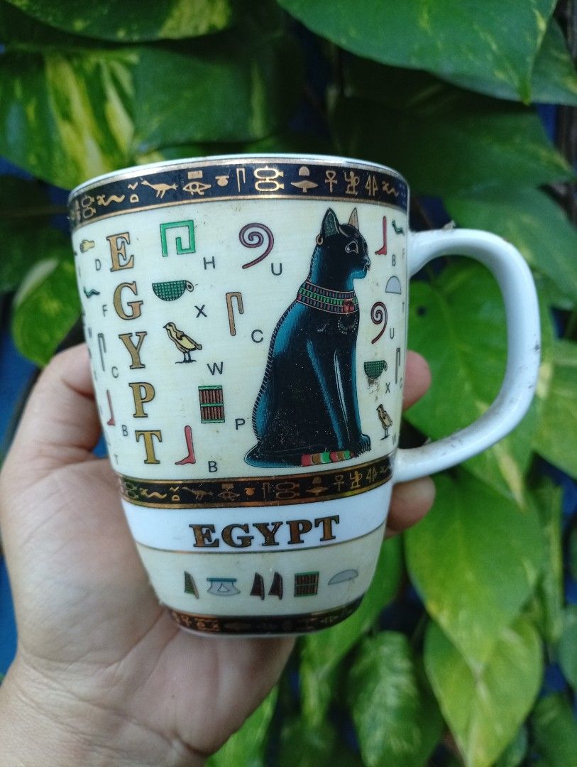 Egyptian Cat Mug with Hieroglyphs (Category: Coffee & Tea Tableware) located in Bacolod City. Submitted by closetcleanupbcd (Listing ID 1459650935, Image 1). Description: - White ceramic mug with an Egyptian theme. - Features a black cat and hieroglyphic symbols. - 'EGYPT' text prominently displayed on the front. - Maker's mark on the bottom includes a camel design and 'Made in Egypt'. - Handle has gold trim, showing some signs of wear. - Minor i