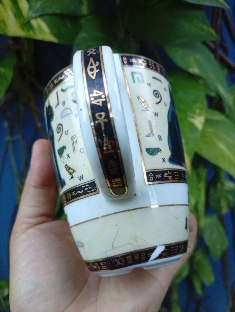 Egyptian Cat Mug with Hieroglyphs (Category: Coffee & Tea Tableware) located in Bacolod City. Submitted by closetcleanupbcd (Listing ID 1459650935, Image 3). Description: - White ceramic mug with an Egyptian theme. - Features a black cat and hieroglyphic symbols. - 'EGYPT' text prominently displayed on the front. - Maker's mark on the bottom includes a camel design and 'Made in Egypt'. - Handle has gold trim, showing some signs of wear. - Minor i