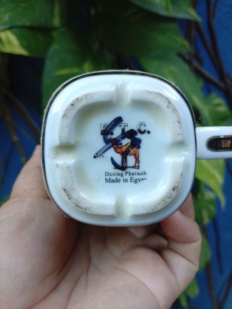 Egyptian Cat Mug with Hieroglyphs (Category: Coffee & Tea Tableware) located in Bacolod City. Submitted by closetcleanupbcd (Listing ID 1459650935, Image 2). Description: - White ceramic mug with an Egyptian theme. - Features a black cat and hieroglyphic symbols. - 'EGYPT' text prominently displayed on the front. - Maker's mark on the bottom includes a camel design and 'Made in Egypt'. - Handle has gold trim, showing some signs of wear. - Minor i