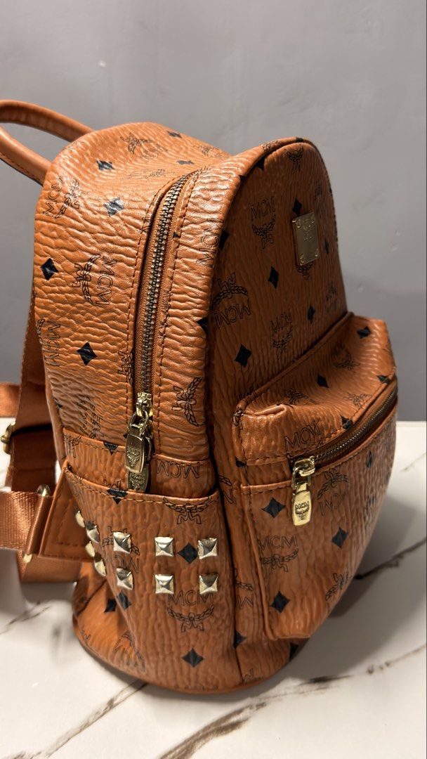 MCM Visetos Mini Backpack Cognac Studded (Category: Backpacks) located in Manila. Submitted by faye_preloved (Listing ID 1459604250, Image 2). Description: - Authentic MCM mini backpack in cognac Visetos - Iconic all-over MCM logo print - Gold-tone hardware and stud details on sides - Adjustable shoulder straps - Top handle - Front zip pocket - Interior pocket - Ideal for everyday use.