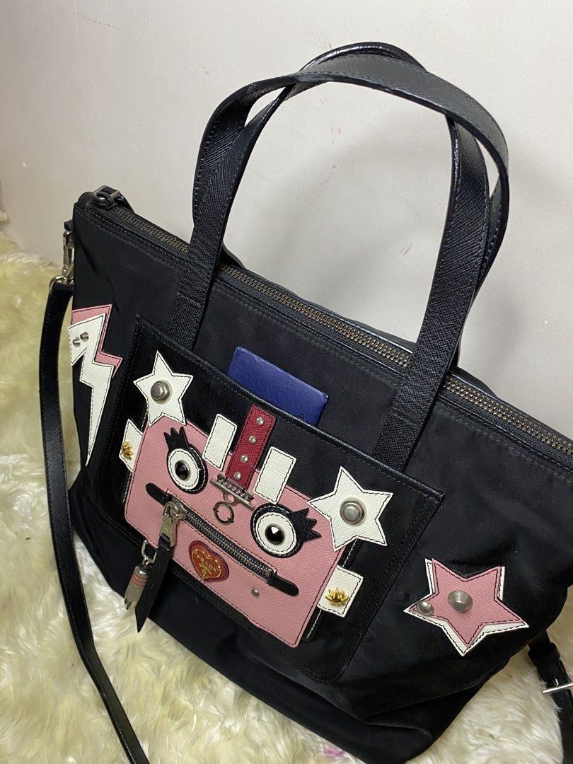 Prada Nylon Robot Applique Tote Bag (Category: Luxury Bags & Wallets) located in Manila. Submitted by ichiban_finds (Listing ID 1459592202, Image 3). Description: - Authentic Prada black nylon tote bag - Features a charming pink robot face applique with star and lightning bolt details - Comes with a detachable shoulder strap for versatile carrying - Interior lined with signature Prada logo - Authenticity card included - Issue can be seen in the f