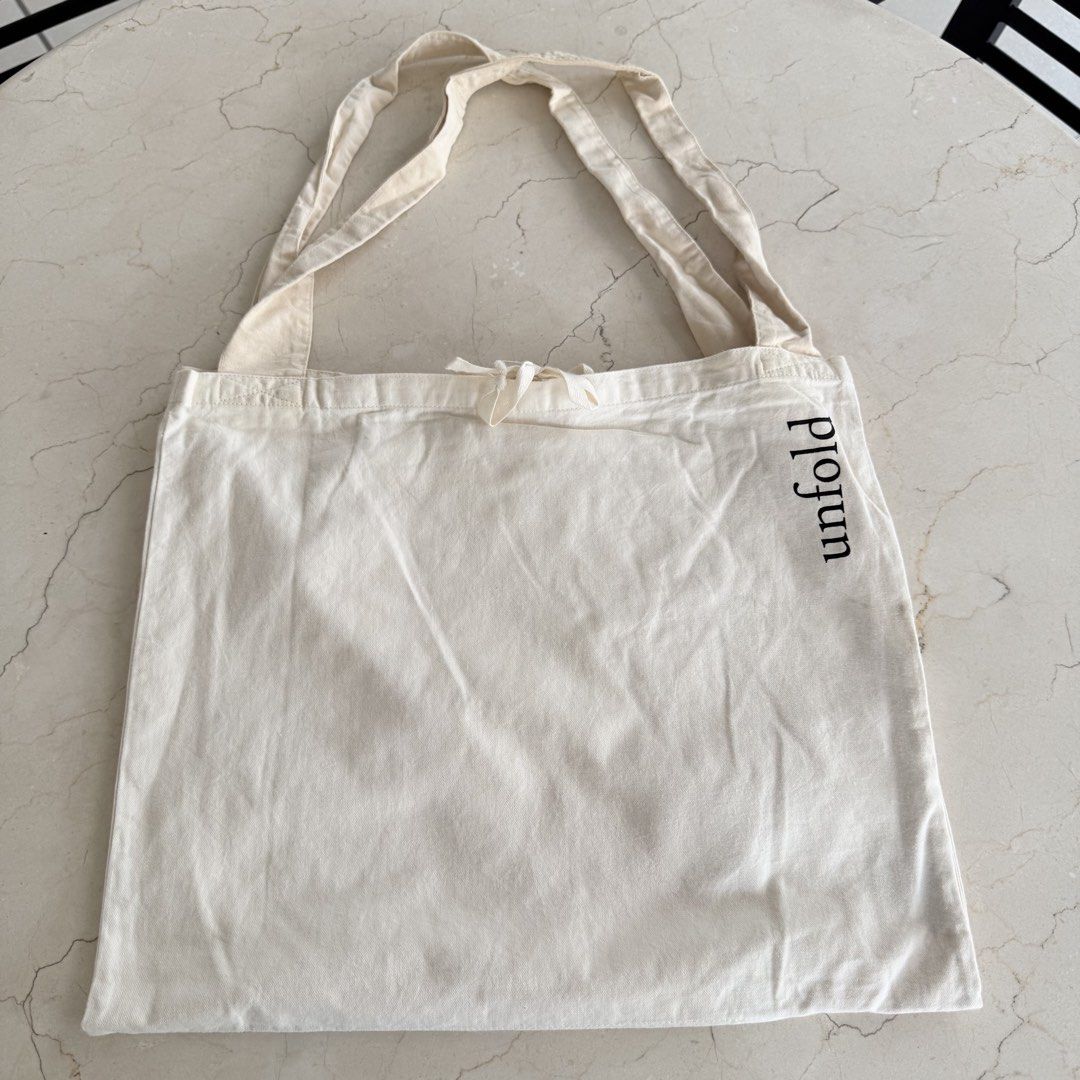 Unfold Canvas Tote Bag (Category: Tote Bags) located in Quezon City. Submitted by lil0.phbyrheadlcrz (Listing ID 1459898920, Image 2). Description: - Cream colored canvas tote bag - 'unfold' printed vertically on the side - Secure drawstring closure at the top - Interior pocket for small items - Ideal for everyday use, groceries, or as a casual carry-all - Simple and functional design.