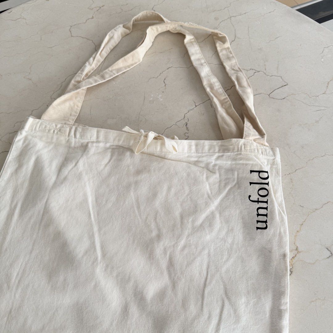 Unfold Canvas Tote Bag (Category: Tote Bags) located in Quezon City. Submitted by lil0.phbyrheadlcrz (Listing ID 1459898920, Image 3). Description: - Cream colored canvas tote bag - 'unfold' printed vertically on the side - Secure drawstring closure at the top - Interior pocket for small items - Ideal for everyday use, groceries, or as a casual carry-all - Simple and functional design.