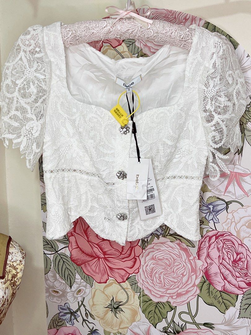 Elegant Bebe White Lace Crop Blouse (Category: Blouses) located in Cavite. Submitted by wishie_collects (Listing ID 1459946761, Image 1). Description: It features a structured, romantic design with a square neckline, delicate lace puff sleeves, rhinestone-embellished decorative front buttons, and a scalloped hem..