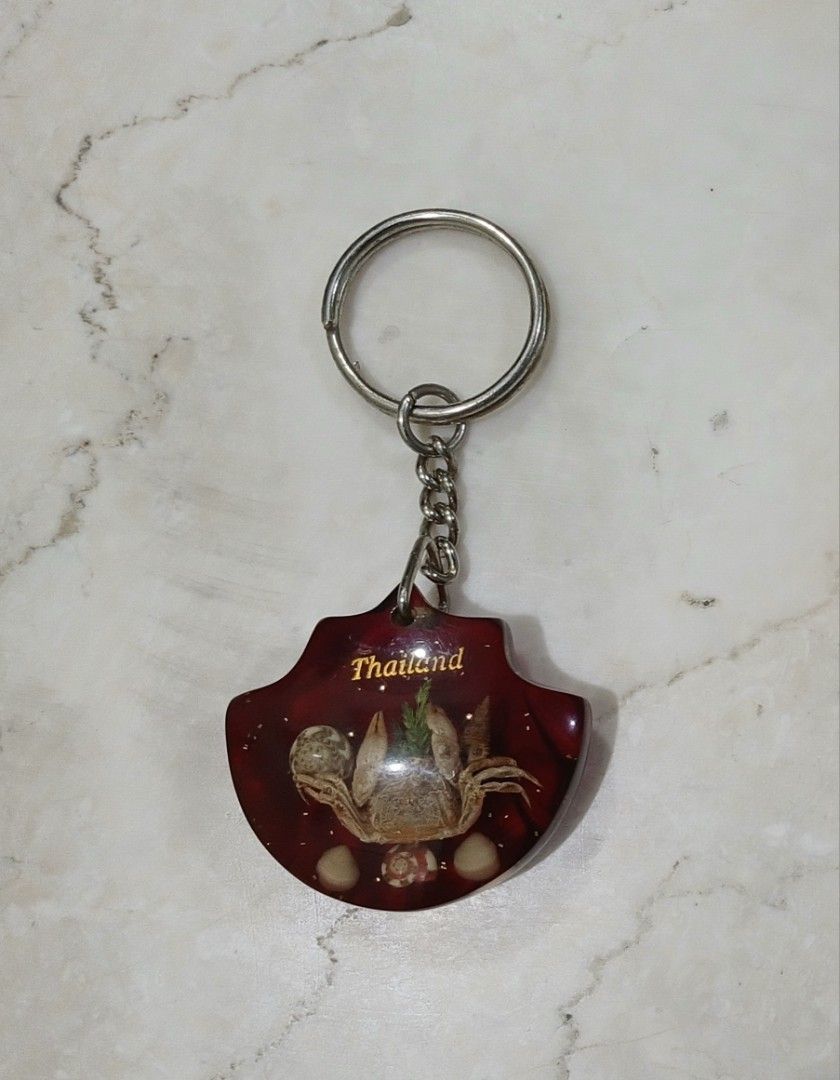 Thailand Souvenir Crab & Seashell Resin Keychain (Category: Travel Essentials & Accessories) located in Bulacan. Submitted by honhonliz (Listing ID 1460025549, Image 1). Description: Vintage souvenir keychain from Thailand. It features a real miniature crab, natural seashells, and small green decorative accents permanently encased in a glossy, fan-shaped deep red translucent epoxy resin pendant. The country name "Thailand" is neatly stamped in go