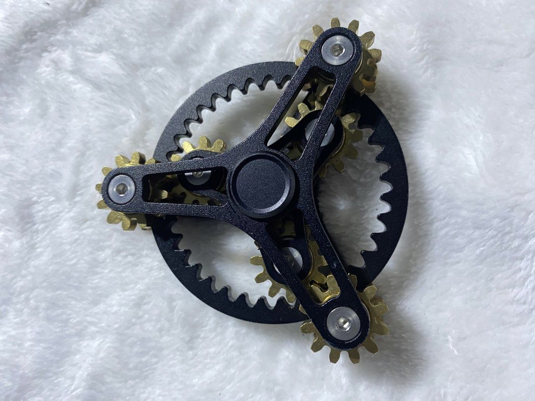 Gear Linkage Fidget Spinner (Category: Action & Battle Toys) located in Iba. Submitted by leovincee.1414 (Listing ID 1460638584, Image 1). Description: Design: A black metallic tri-arm frame with interlocking brass-colored cogs and gears. Materials: Typically made with an aluminum or alloy body and solid brass gears. Function: Designed for stress relief, anxiety reduction, and tactile focus by spinning and turning the meshed gear system. - Time, 