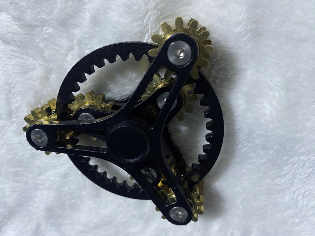 Gear Linkage Fidget Spinner (Category: Action & Battle Toys) located in Iba. Submitted by leovincee.1414 (Listing ID 1460638584, Image 3). Description: Design: A black metallic tri-arm frame with interlocking brass-colored cogs and gears. Materials: Typically made with an aluminum or alloy body and solid brass gears. Function: Designed for stress relief, anxiety reduction, and tactile focus by spinning and turning the meshed gear system. - Time, 
